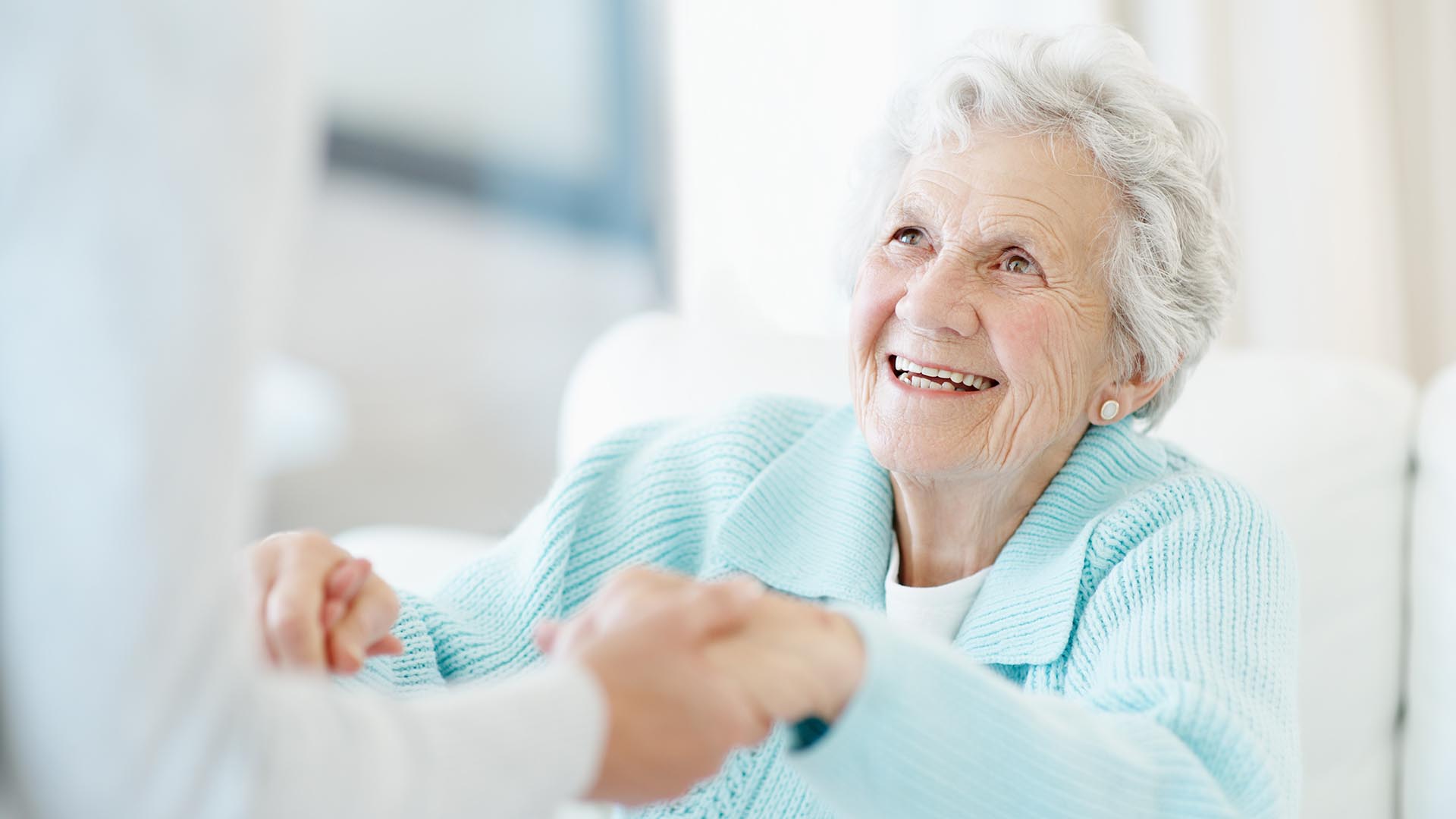 Alter Damen mit weißen Haaren und hellblauem Pullover, die freudig strahlt. Ihr werden helfende Hände gereicht, um einfacher aufstehen zu können.